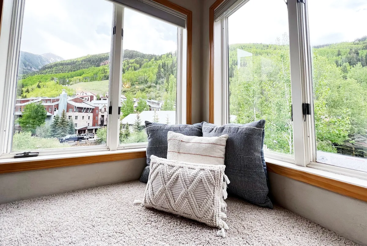 A serene living area with large windows that allow natural light to pour in, plush pillows on a comfortable seating area, and a carpeted floor. The windows frame a stunning view, creating a seamless connection between the interior and the scenic outdoors.
