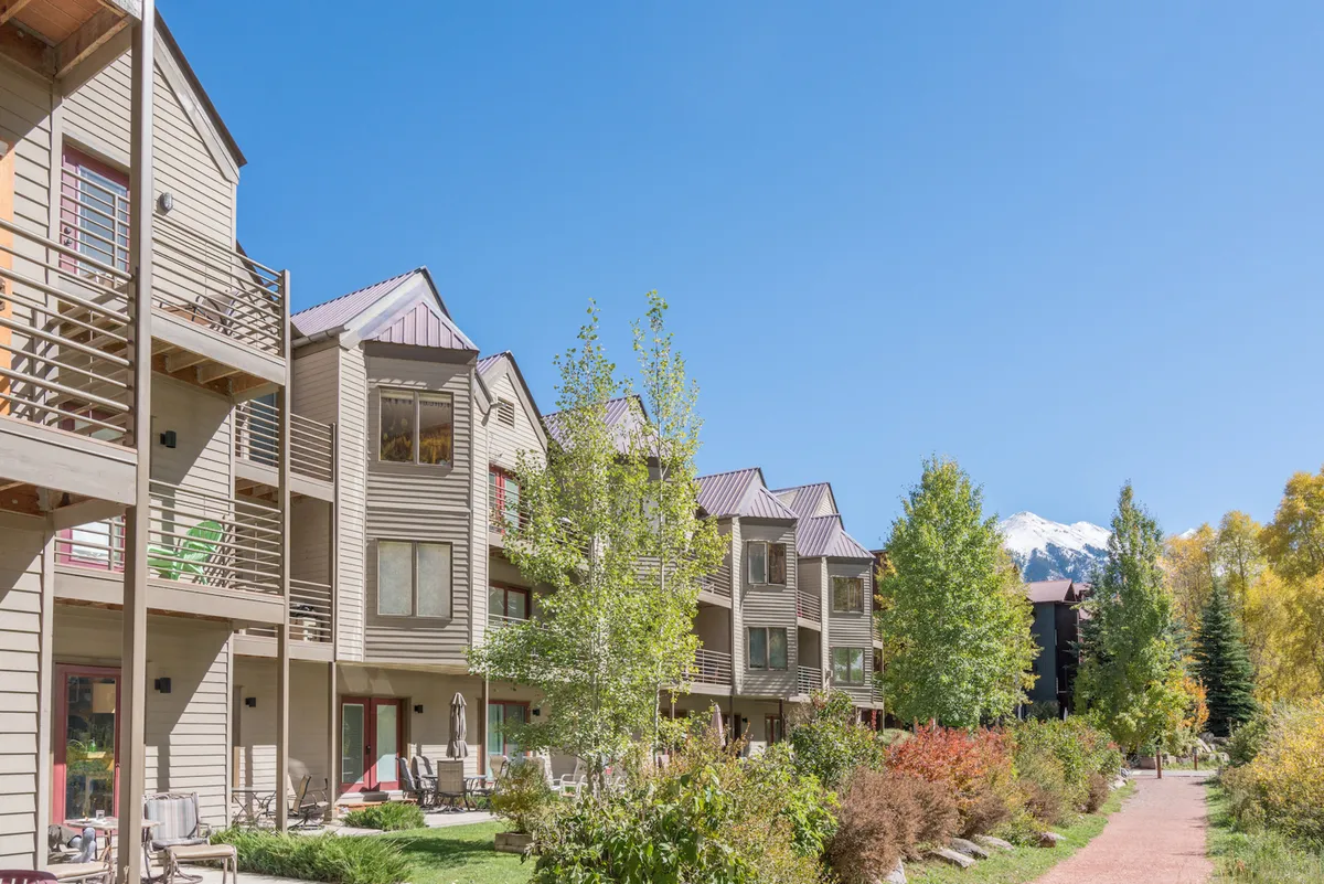 The image captures a modern exterior with landscaped gardens and a striking mountain backdrop. Residents relax on balconies or stroll through the garden, enjoying mountain views and the peaceful environment.