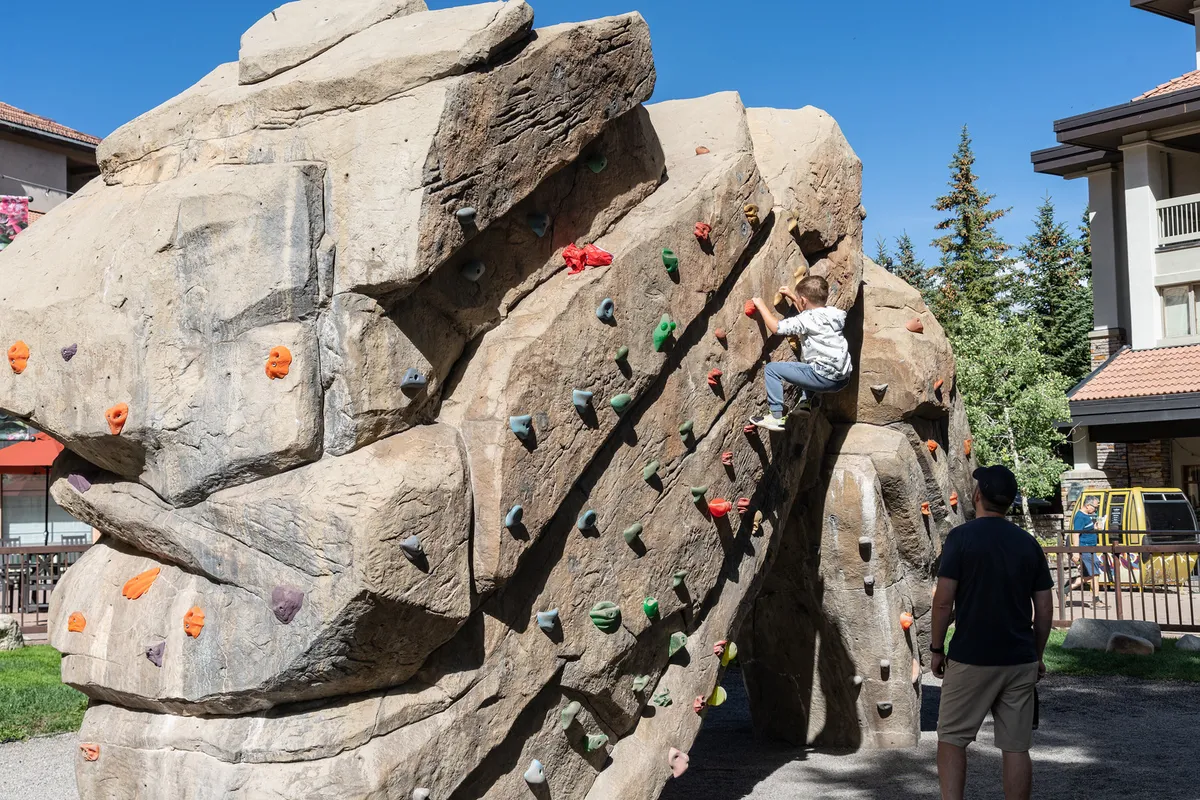 A lively exterior scene at a local park featuring a child confidently scaling a rock climbing wall, closely supervised by an adult. The background includes lush greenery and play equipment, emphasizing active, outdoor fun in a safe environment.