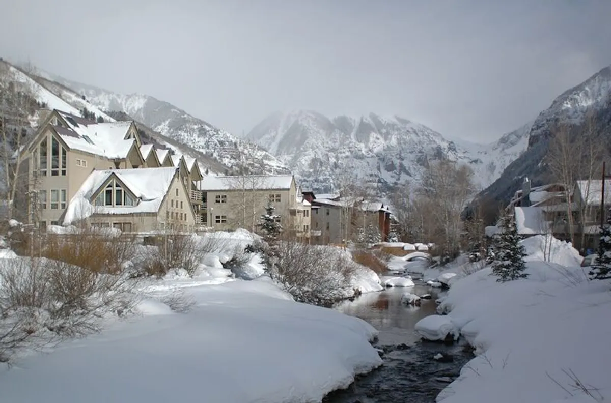 A sweeping winter scene shows snow-covered mountains, a flowing stream, snow-laden trees, and nearby buildings. The peaceful atmosphere and natural beauty evoke both adventure and tranquility, ideal for couples seeking outdoor thrills and romantic strolls.