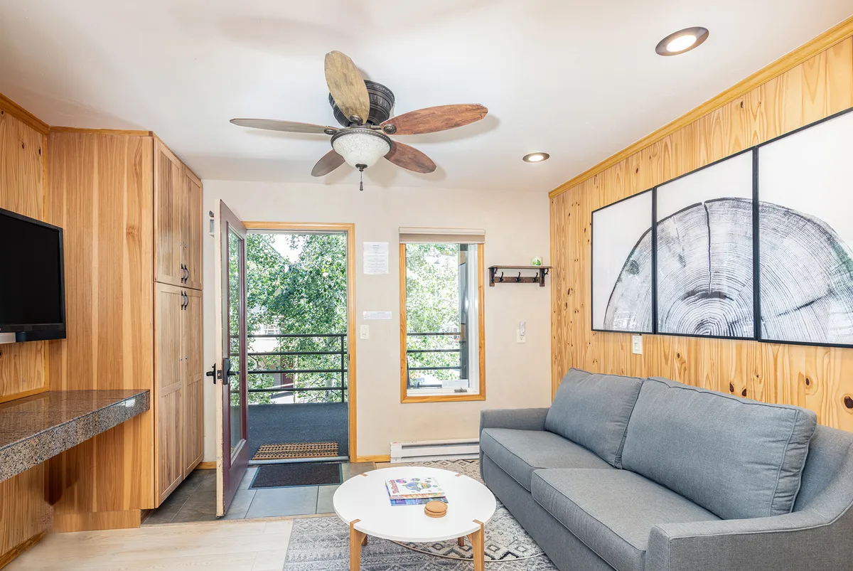 The image showcases a warm, sunlit living area with a plush gray sofa, rustic wooden accent wall, stylish ceiling fan, and a coffee table, all set beside expansive windows that frame views of the surrounding mountains and river. The inviting layout is perfect for relaxing, reading, or gathering with friends, while abundant natural light highlights the cozy ambiance.