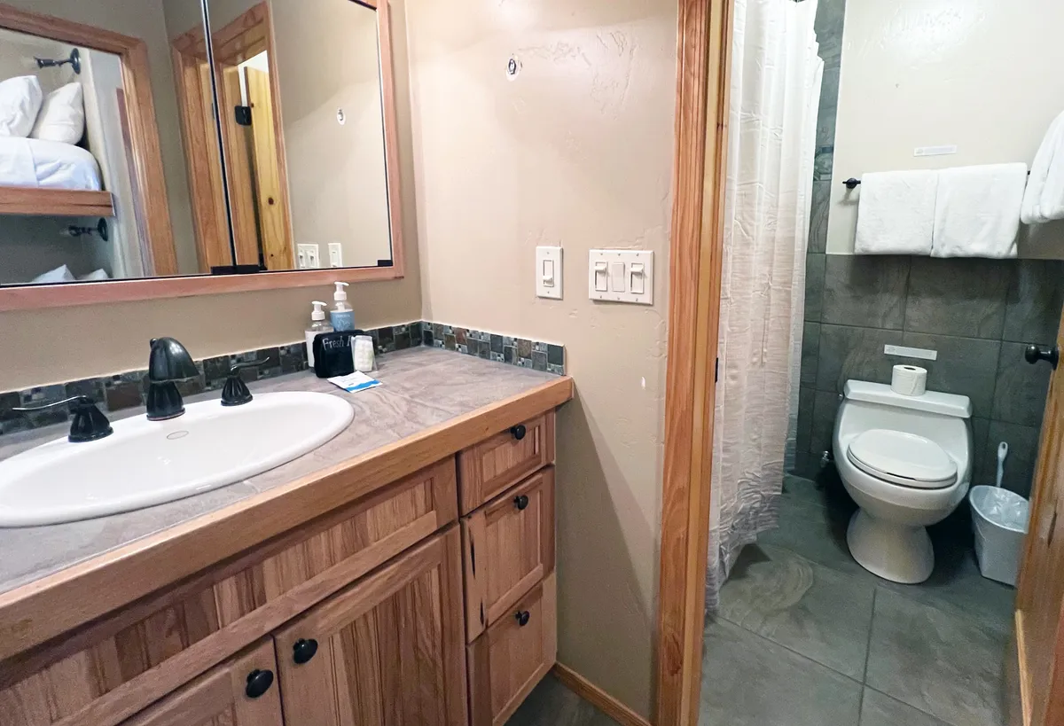 This image shows a spacious bathroom featuring a sink with a large mirror, a toilet, a shower curtain, and wooden cabinetry. The inviting and functional space is well-lit and features warm wood tones that contribute to a cozy atmosphere. This view highlights the bathroom's practicality while suggesting comfort and relaxation.