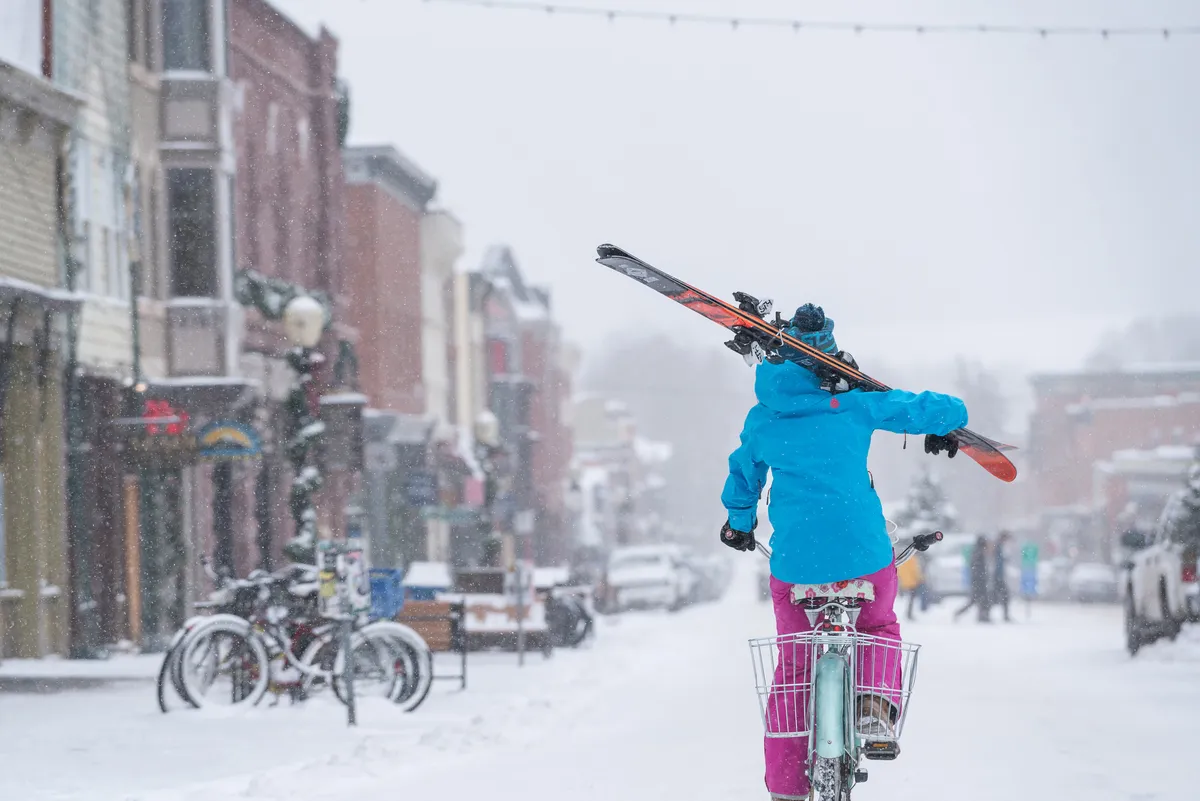 A lively street scene with a person biking while carrying skis, colorful storefronts, and snow-laden sidewalks. The vibrant town atmosphere is evident, showcasing locals and visitors exploring shops and enjoying the lively winter landscape.