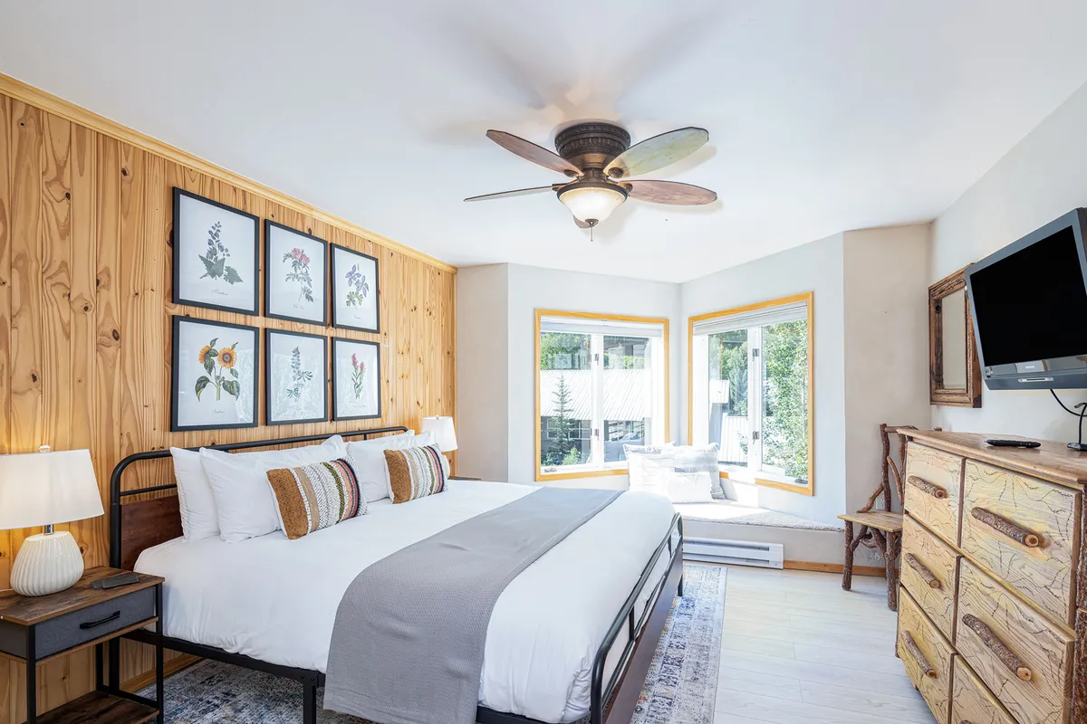 A king bed with decorative pillows is positioned near large windows that let in abundant natural light. Framed botanical prints and a rustic dresser add warmth and character to the space, while the overall ambiance feels serene and inviting for parents.