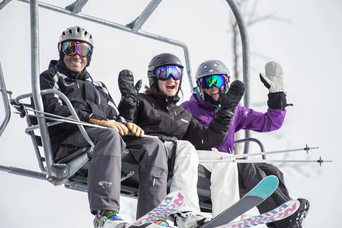 Three skiers ride a chairlift above a snow-covered landscape, equipped with ski gear and surrounded by stunning mountain views. The mood is lively and adventurous, highlighting the excitement of winter sports right outside the doorstep.