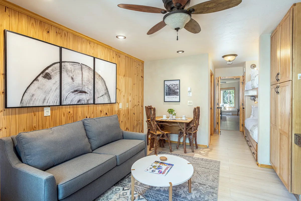 A compact interior space featuring a sofa, a dining table, wooden wall art, and a decorative round table. Morning sunlight filters in through the windows, creating a homely and connected feel, ideal for small family gatherings or shared daily routines.