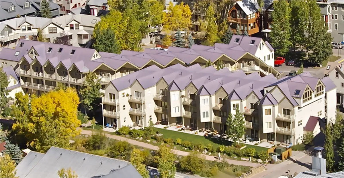 Rows of welcoming townhouses set amidst landscaped gardens and towering trees, with outdoor dining and gathering spaces visible. The setting hints at friendly communal spaces perfect for BBQs and summer gatherings.