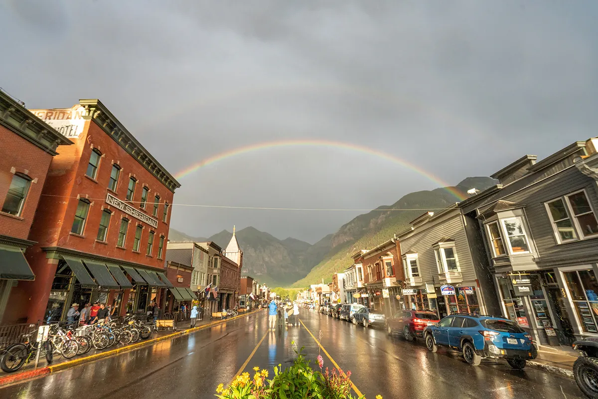 A vibrant street scene with historic buildings, a colorful rainbow arching overhead, wet pavement, and bicycles parked along the sidewalk. This lively setting conveys Telluride’s inviting town center, perfect for shopping, dining, and exploration, in any season.
